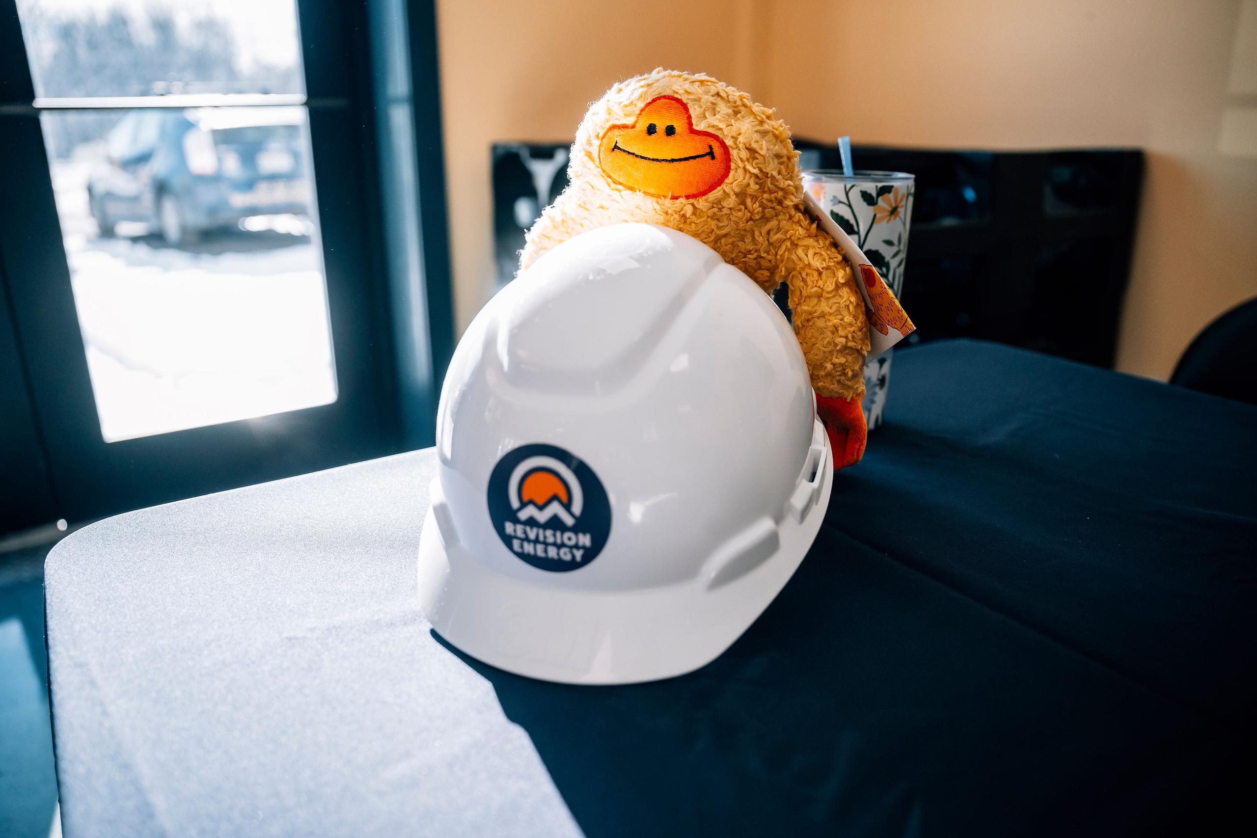 Sunsquatch plushy behind a ReVision Energy hardhat